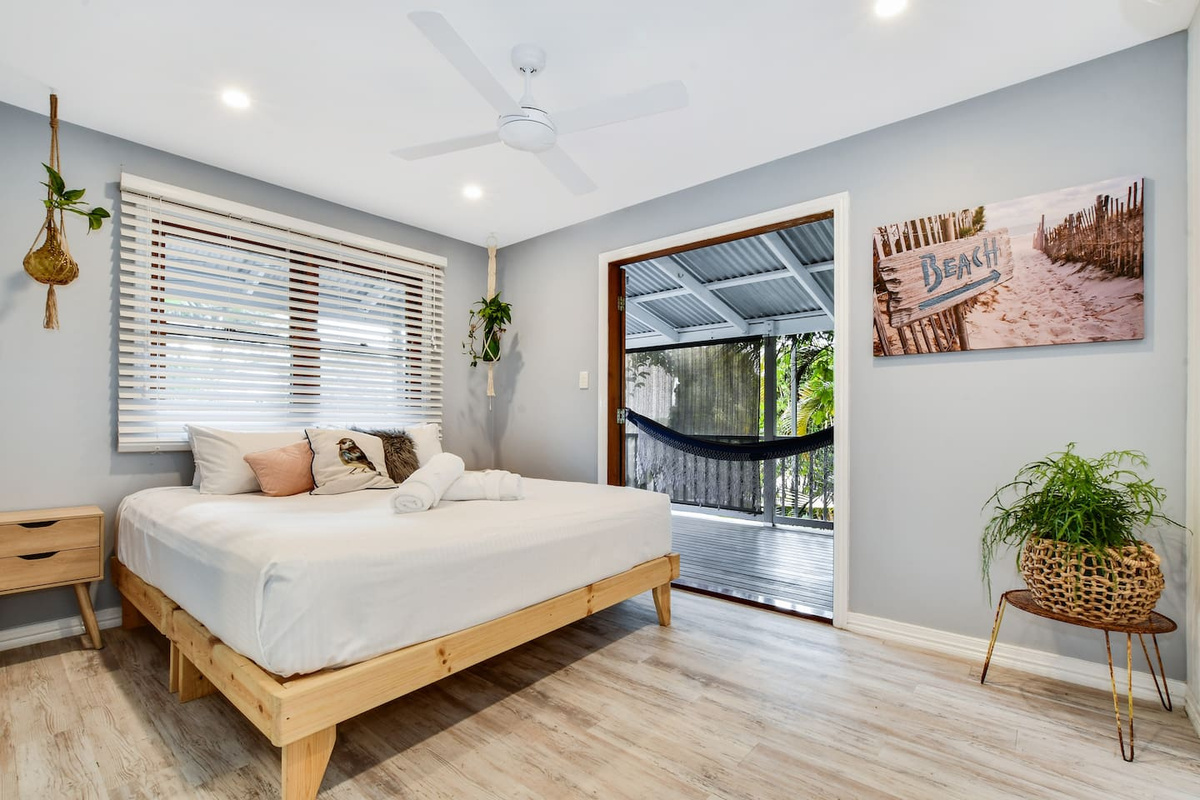 Timber-framed openings, light coastal finishes and direct outdoor access give this queen bedroom at Aaron's Beachside Boat House plenty of beach-house character.