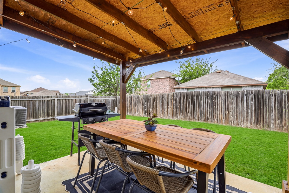 Covered backyard patio with wood dining table, string lights, and propane BBQ grill — perfect for alfresco meals and evening cookouts. Fully fenced for privacy with direct access from the kitchen.