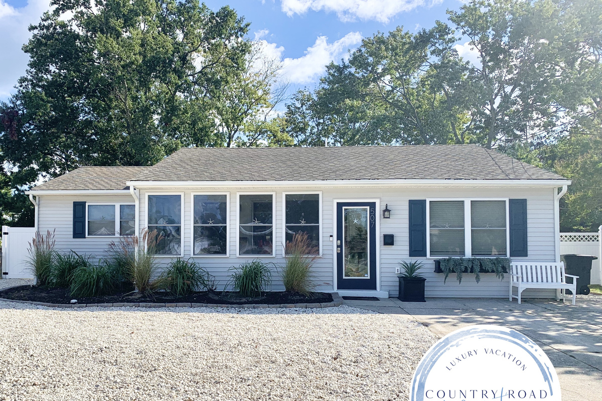 Welcome to your getaway! This lovely ranch-style home features light gray siding, navy shutters, and a bright sunroom. The exterior is complete with a white stone and manicured greenery, offering great curb appeal and a peaceful setting.