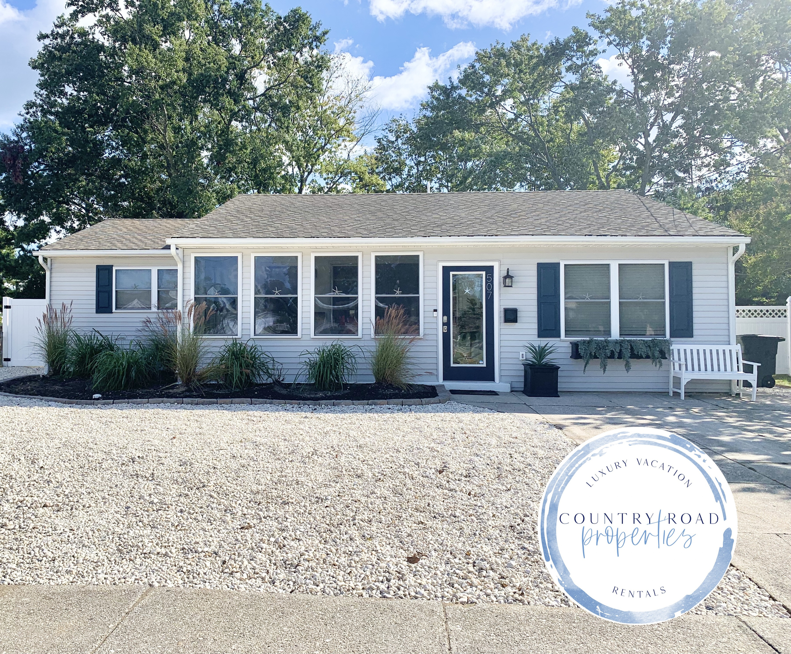 Welcome to your getaway! This lovely ranch-style home features light gray siding, navy shutters, and a bright sunroom. The exterior is complete with a white stone and manicured greenery, offering great curb appeal and a peaceful setting.