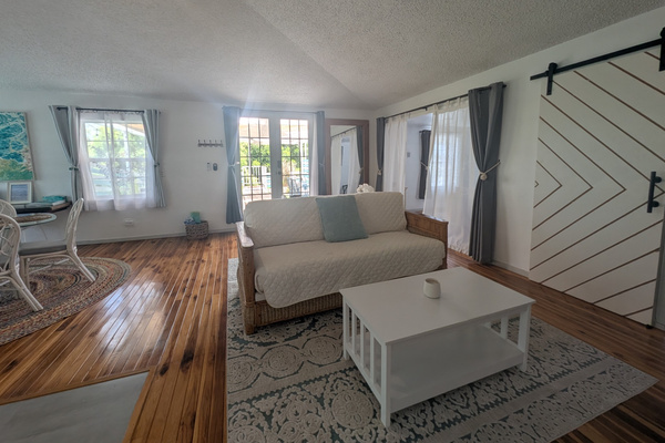 Upstairs living room at Villa Coral featuring a white couch, matching coffee table, patterned area rug, chevron barn door, and glass French doors leading out to a private rooftop patio in Dorado, Puerto Rico.
