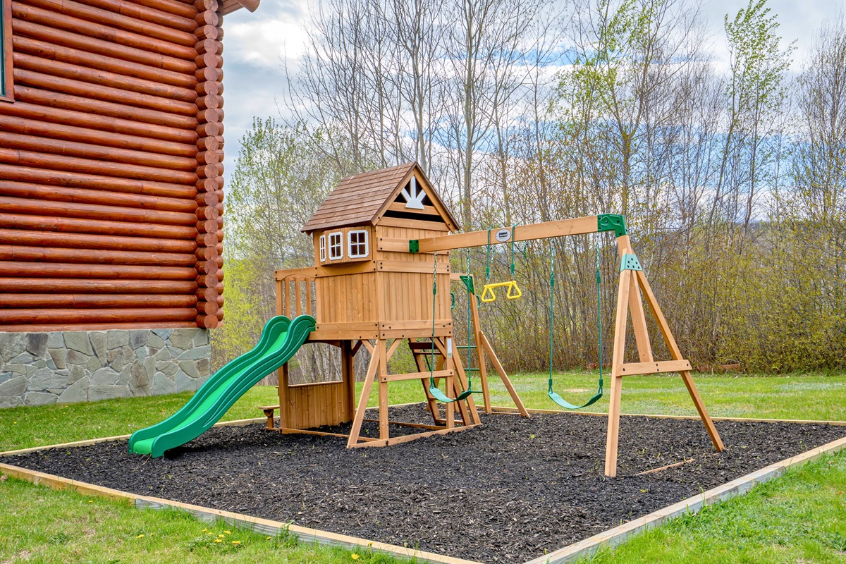 Enjoy family-friendly outdoor fun with this charming wooden playset featuring swings, a slide, and plenty of open green space surrounded by nature.