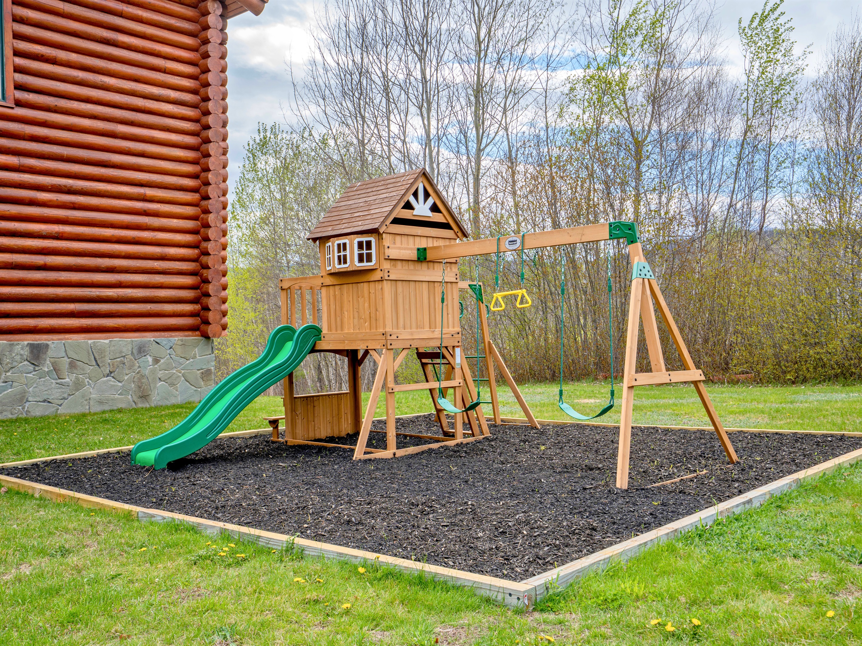 Enjoy family-friendly outdoor fun with this charming wooden playset featuring swings, a slide, and plenty of open green space surrounded by nature.