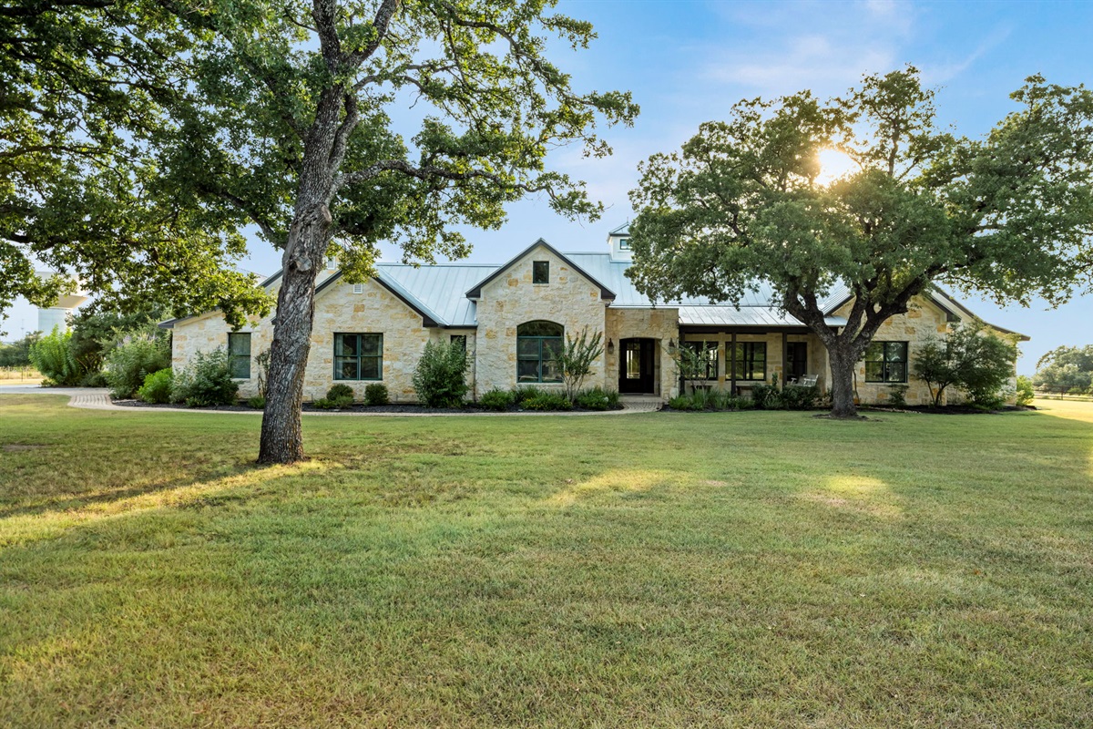 A stone exterior, expansive green lawn, and majestic oak trees under a clear sky.