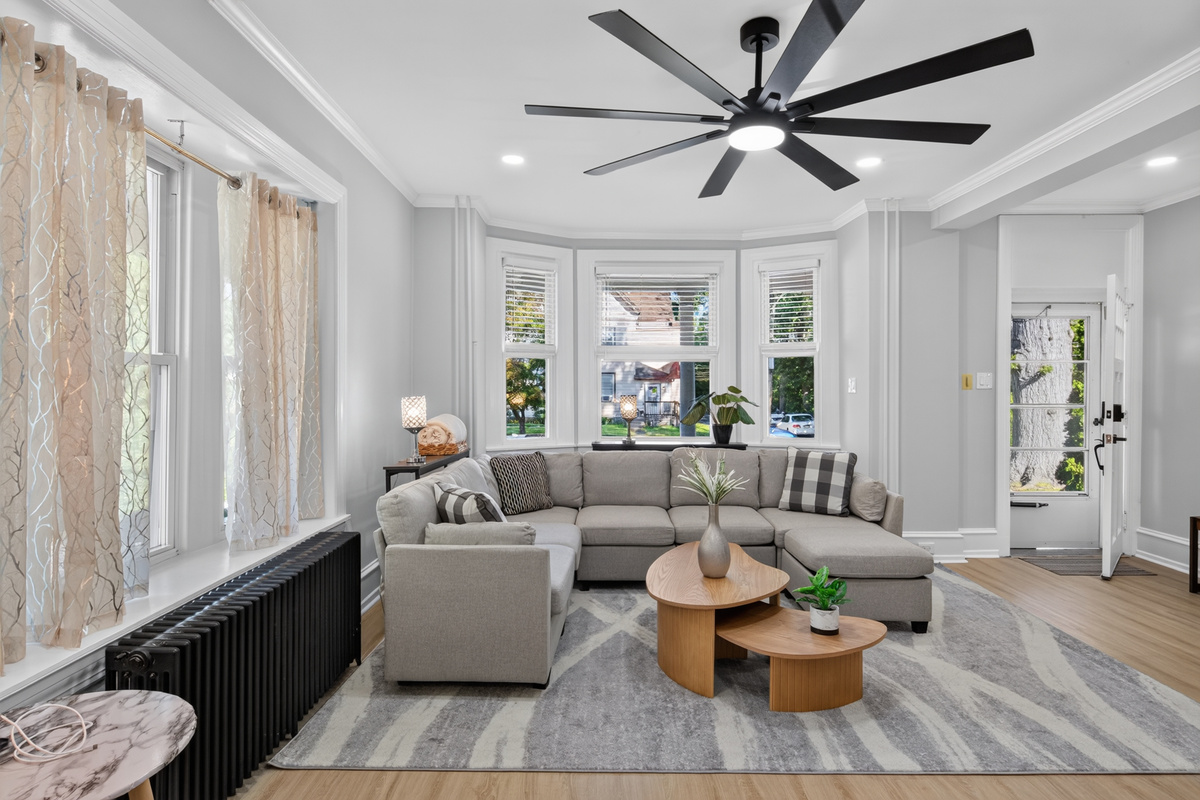 Sunlit open-concept living area with plush sectional seating, ceiling fan, and classic bay windows.