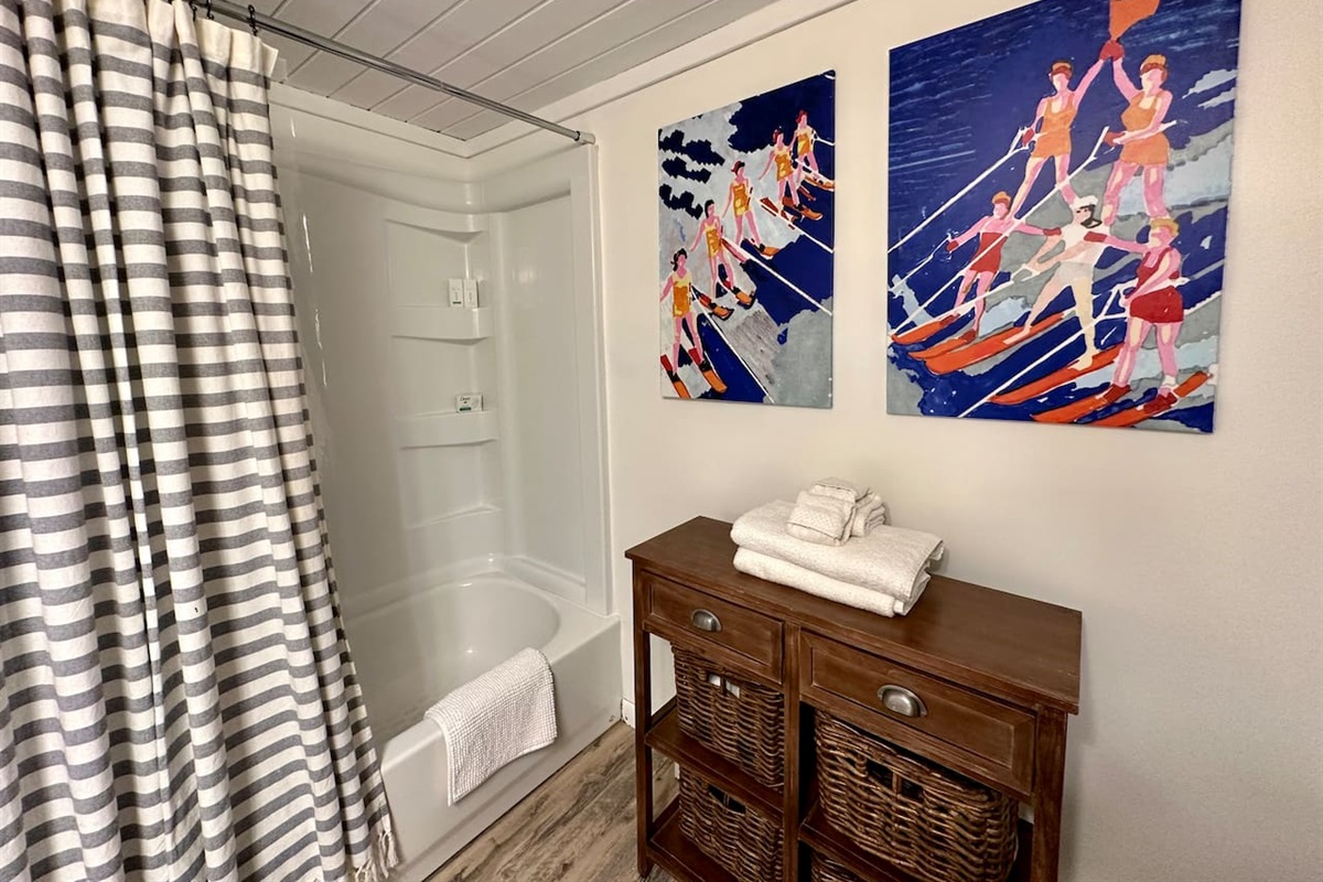 Fresh, coastal-inspired bathroom with a full tub/shower, striped curtain, soft towels, and fun vintage waterski art. A wood cabinet with woven baskets adds charm and storage, making this space both functional and inviting for you.