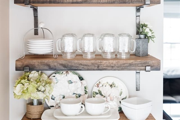 These charming rustic floating shelves are fully stocked with essentials, including mason jar mugs, decorative floral plates, and clean white bowls. Everything is neatly organized and easily accessible for your dining needs.