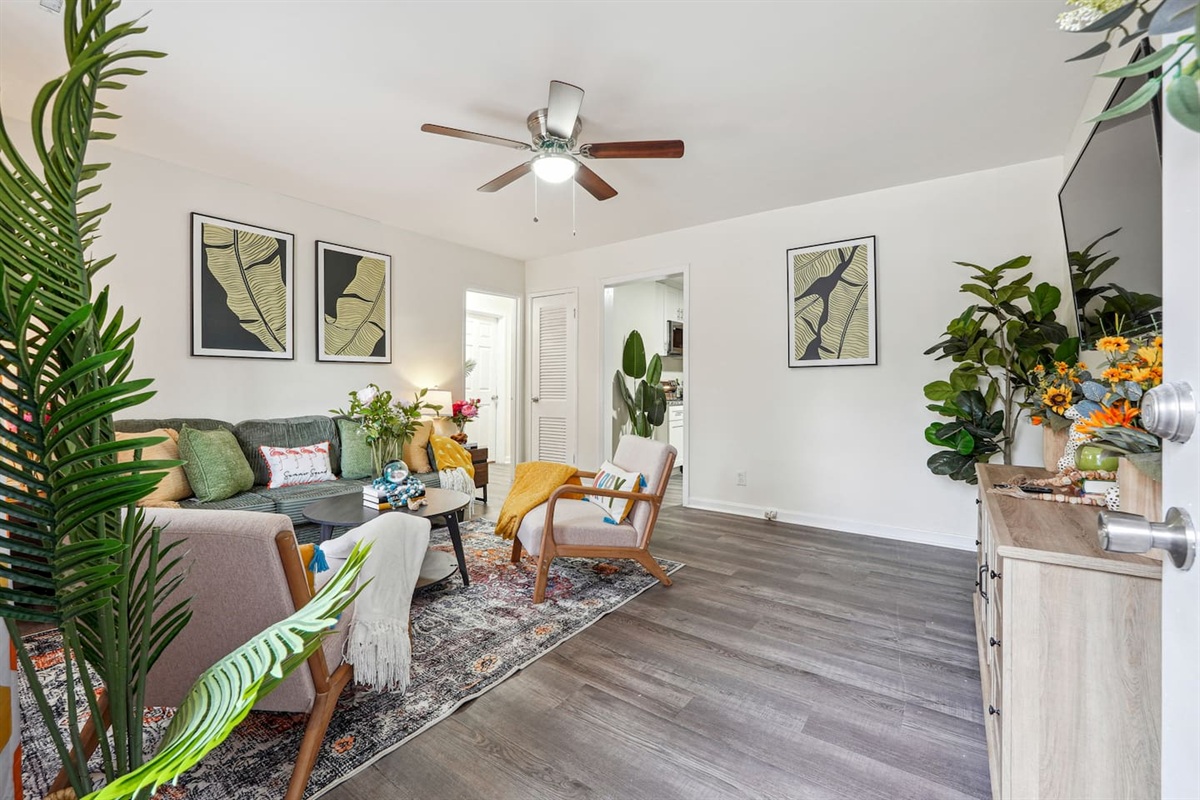 Enjoy a cozy living room featuring a long comfortable sofa, a TV for your entertainment, a ceiling fan, two chair, and a window with a lovely view outside — perfect for relaxing and unwinding.
