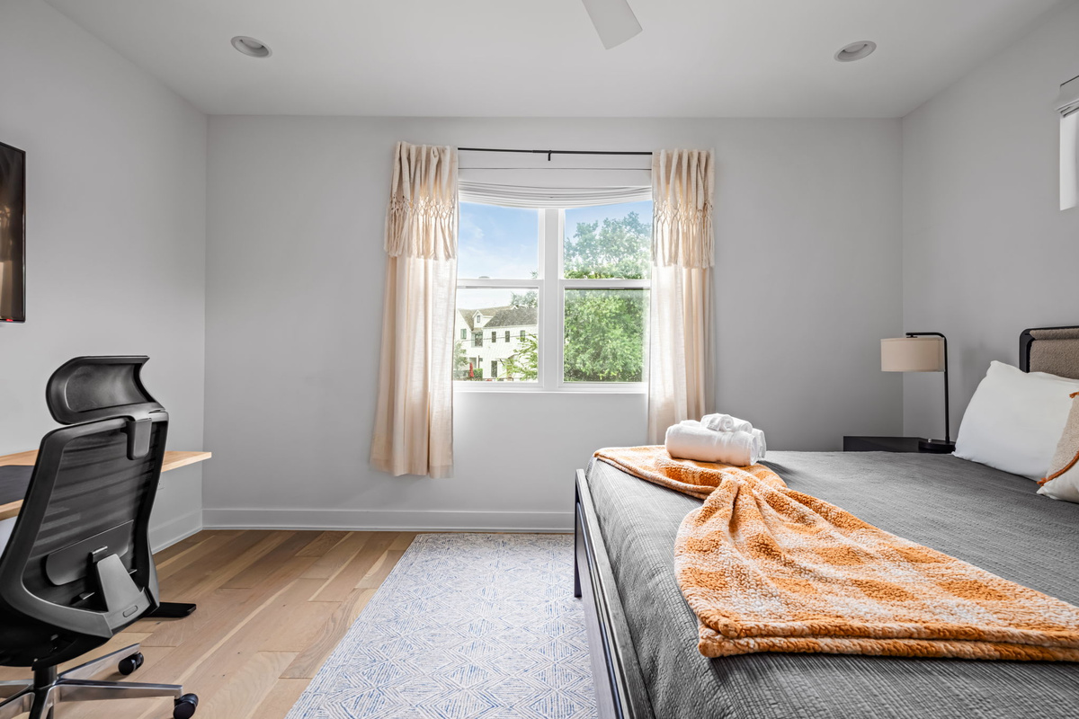 Bright bedroom nook with a comfy daybed, desk chair, and large window overlooking the neighborhood — versatile space for work or rest.
