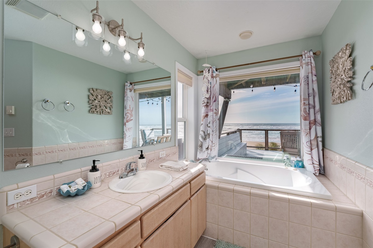 A cozy bathroom with a large window offering a view of a calming beach and the ocean with a corner jacuzzi. 