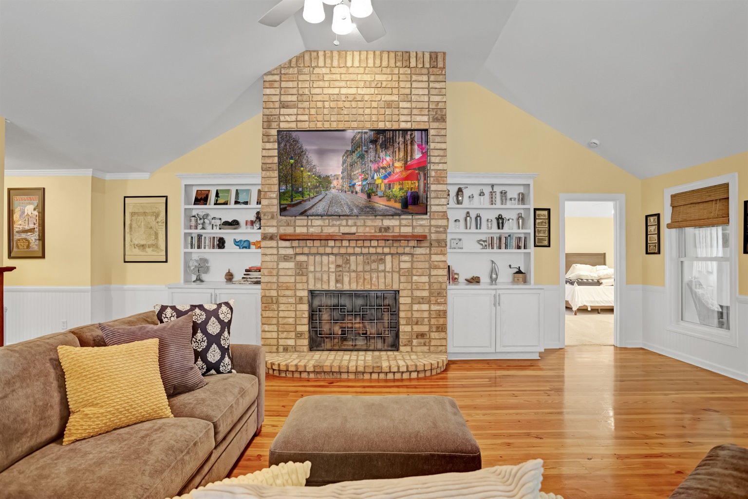 Cozy living room with vaulted ceilings, warm hardwood floors, and a striking brick fireplace centerpiece, flanked by custom built-ins and a mounted flat-screen TV.