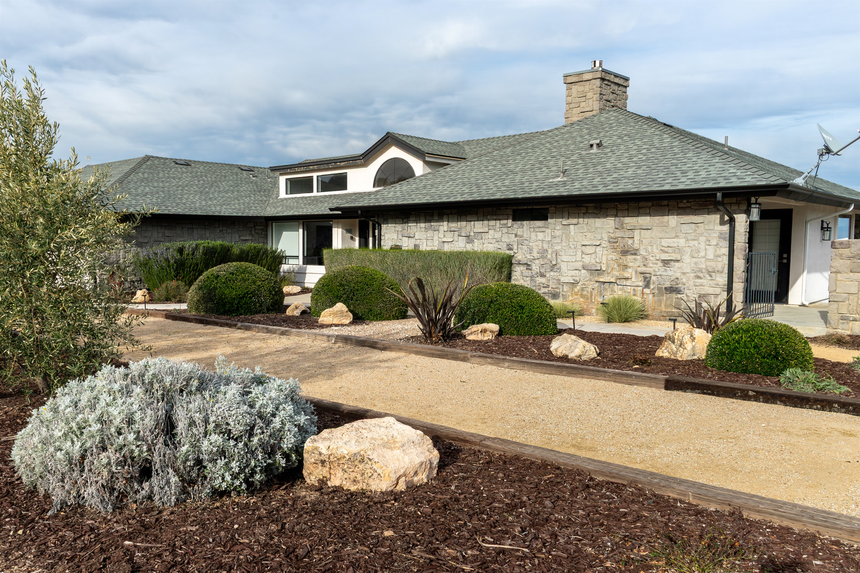 Drought-tolerant landscaping, native stone, and an olive tree frame the arrival experience at the Hilltop Guest House — a home that looks like it was built to belong here.