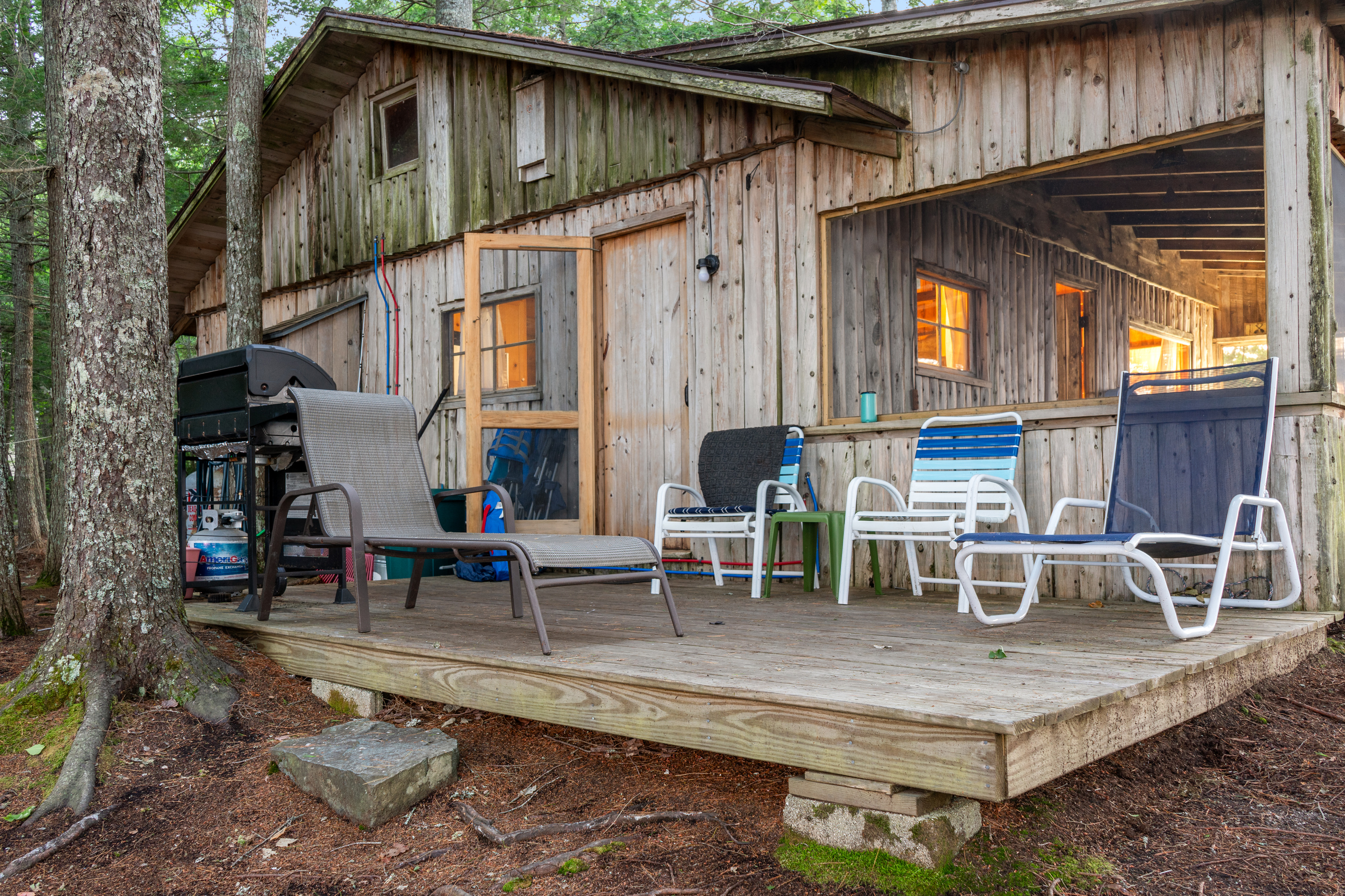 Open wooden deck off the kitchen at Loonrise Point! Simple deck setup on block footings with lawn seating, propane gas grill, and direct door access inside. Weathered cedar siding, exposed PEX lines, and rustic outdoor living on Beech Hill Pond.