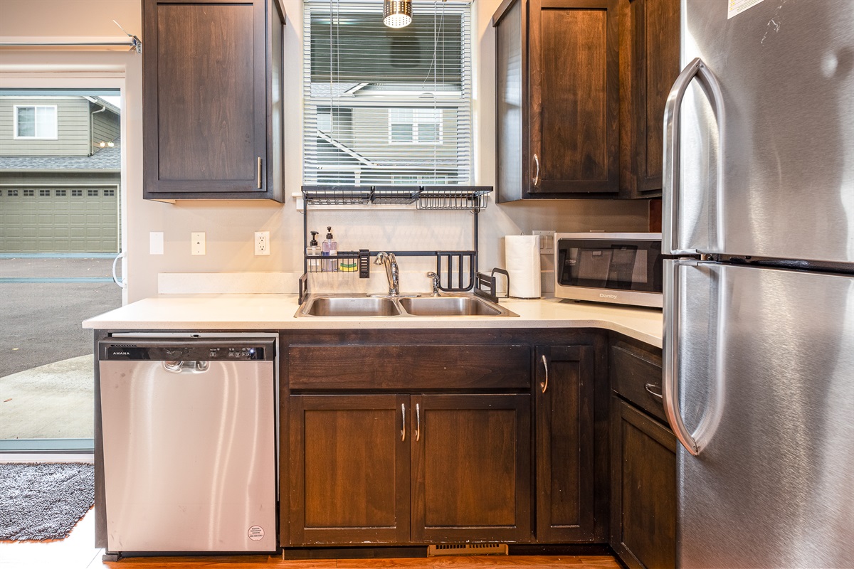 A modern kitchen features dark wooden cabinets, a double sink with a stainless steel faucet, a dishwasher, a microwave, and a window with blinds, all against a backdrop of neutral-colored walls and a view of a garage outside.