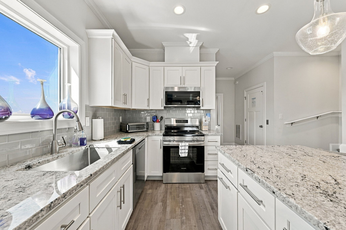 Comprehensive perspective of the chef's kitchen highlighting the expansive granite island, undermount sink, classic white shaker cabinetry, stainless steel range, and soft natural light pouring through the window.   