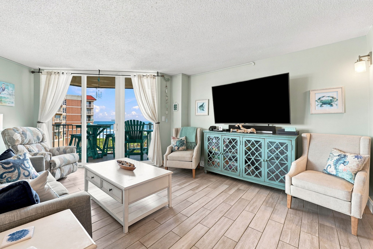 Sun-drenched living area featuring an inviting armchair set, coffee table, and large patio doors open to the coast.