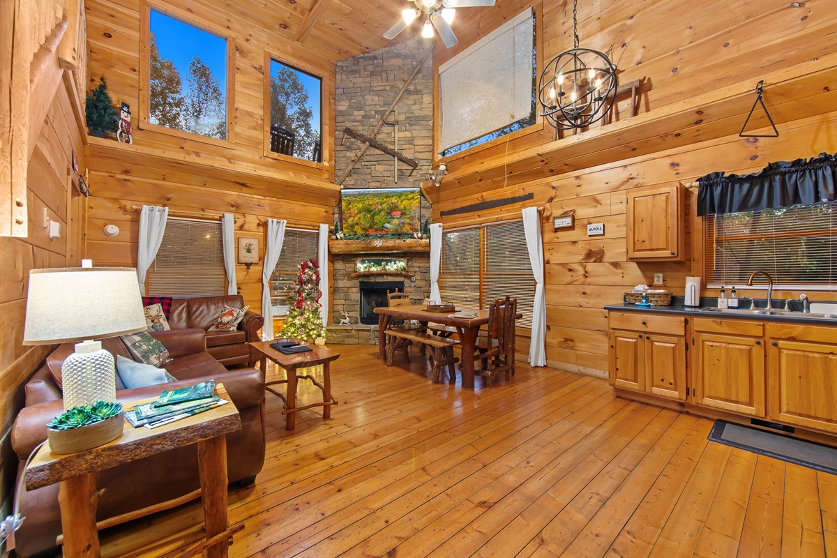 A warm and rustic living space with polished wood floors, a staircase leading to a loft, and an adjacent kitchen with black appliances and dining. The cozy vibe is enhanced by leather couches and charming decor.