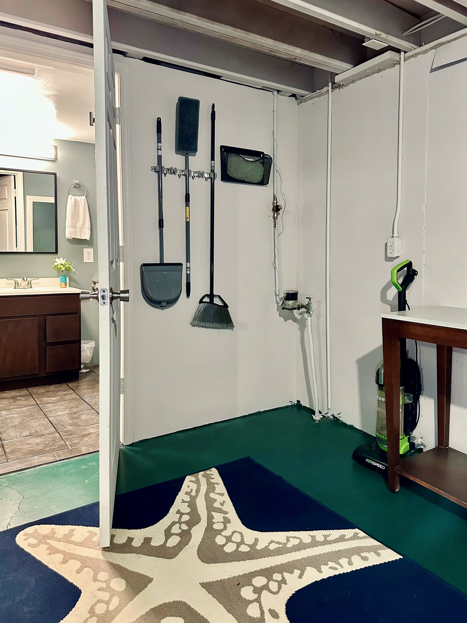 Wall-mounted cleaning station located just outside the basement bathroom. Includes a mop with extra mop pads, broom, dustpan, and vacuum, all neatly stored and easily accessible.