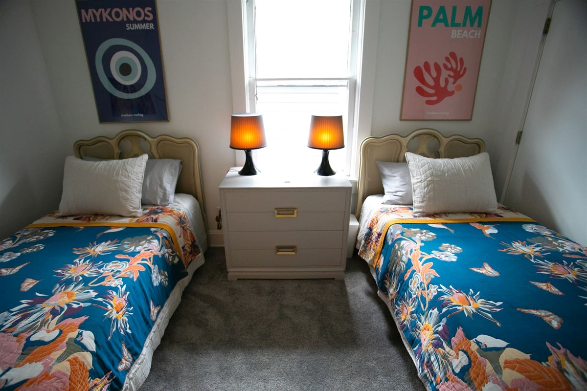 Two twin beds with supportive mattresses, soft cotton linens, blackout shade, ceiling fan & personal bedside lighting — a cozy, sleep-friendly room ideal for shared stays or kids, adjacent to the primary bedroom