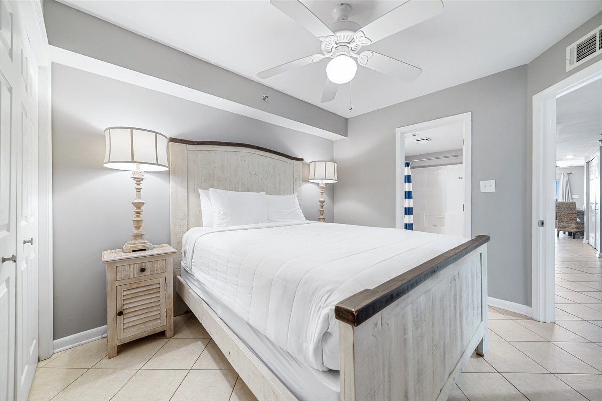 This well-appointed primary bedroom features a king-size bed with a decorative headboard, crisp white bedding, matching nightstands with lamps, and tile flooring for easy maintenance.