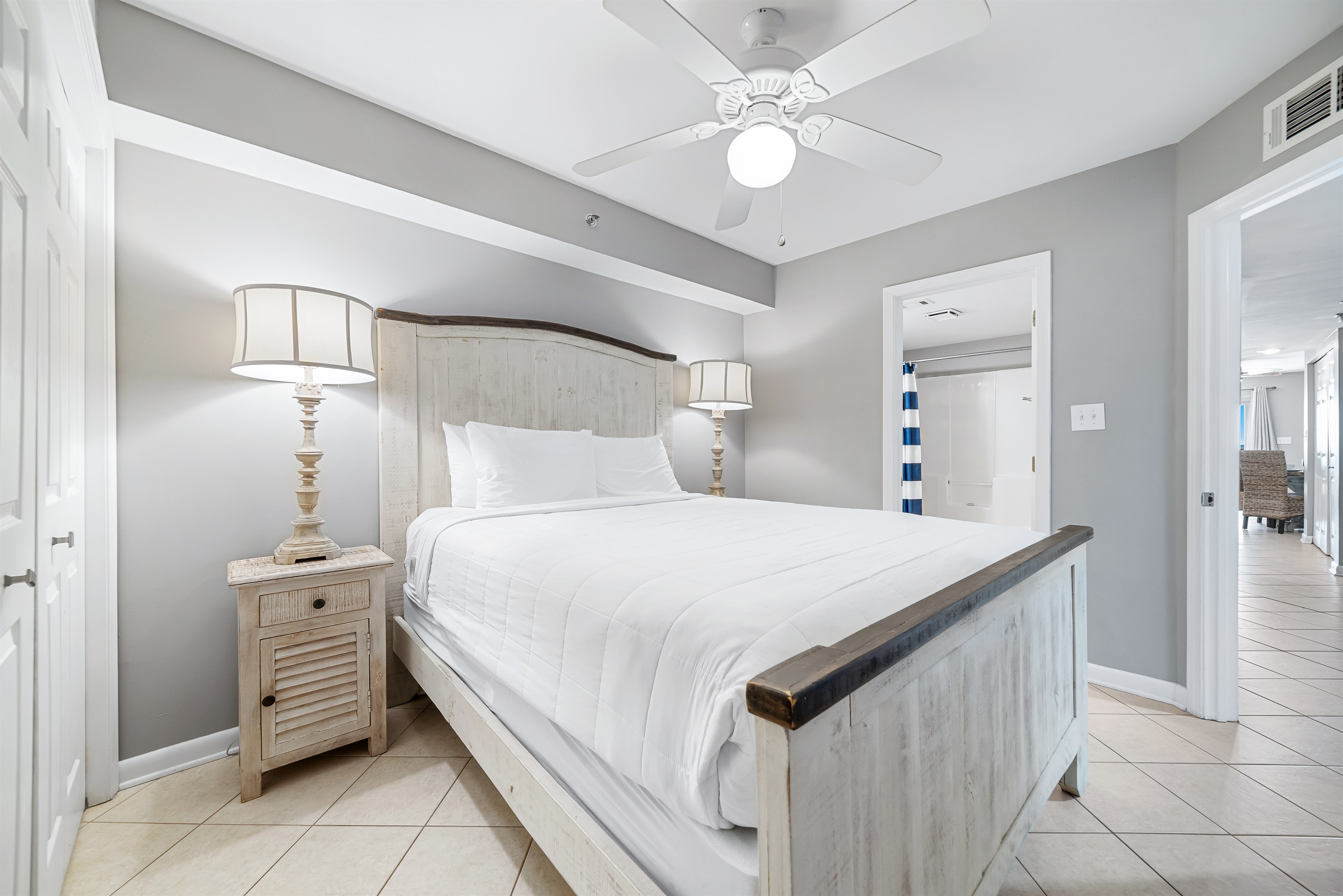 This well-appointed primary bedroom features a king-size bed with a decorative headboard, crisp white bedding, matching nightstands with lamps, and tile flooring for easy maintenance.