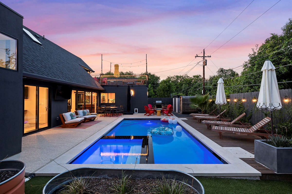 A luxurious backyard at sunset featuring a sparkling swimming pool, a spacious deck with multiple lounge chairs and a dining area, all set against a modern dark gray home with large windows and a rooftop deck.
