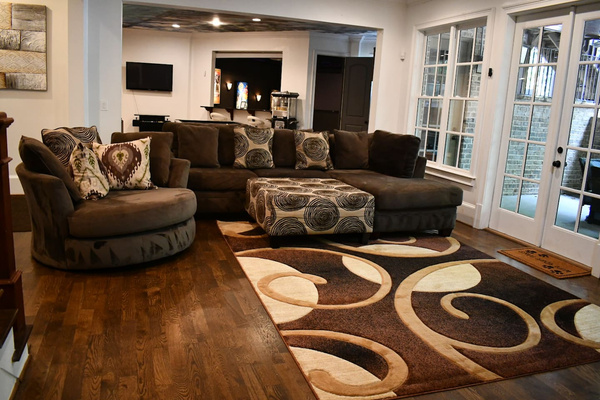 Spacious open-concept living room with hardwood floors, recessed lighting, and crown molding. Features a large sectional sofa with accent pillows, matching ottoman, patterned area rug, and cozy seating arranged for entertaining and relaxation.