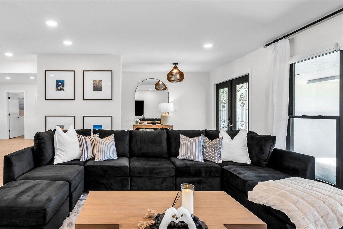 Elegant open-concept living room featuring a plush black velvet sectional, 75” smart TV, and designer mid-century modern furnishings. Floor-to-ceiling windows and French doors fill the space with natural light and connect seamlessly to the outdoor pool.