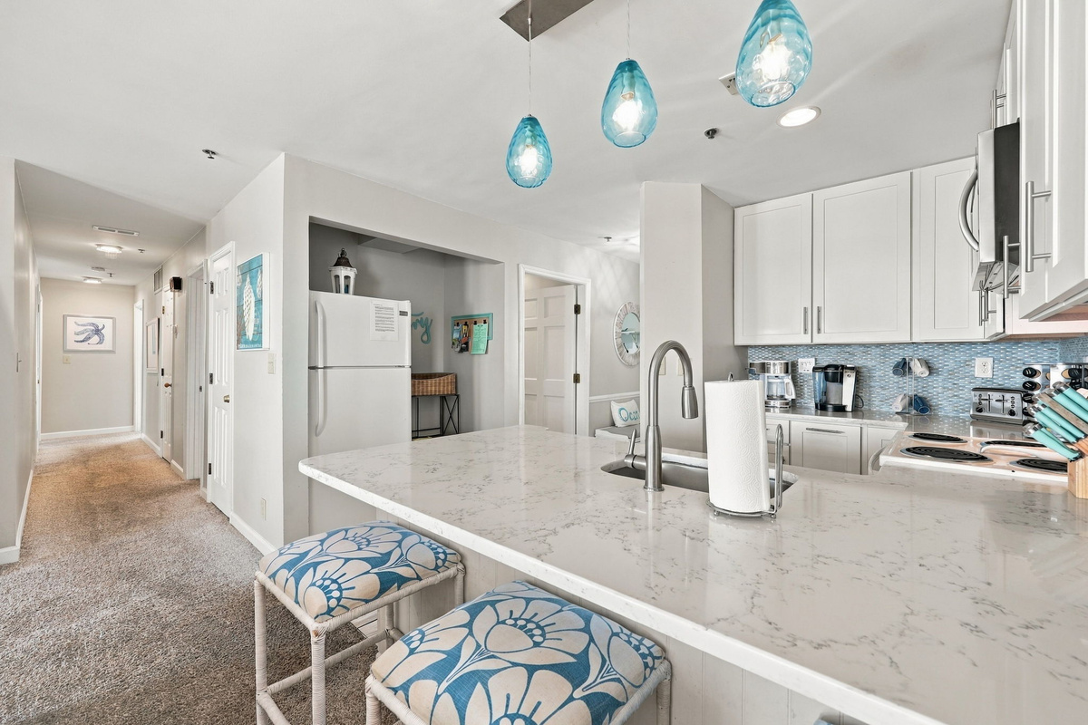 Kitchen island view featuring white veined quartz countertops, undermount sink, gooseneck faucet, blue glass pendant lights, and upholstered barstools overlooking the hallway.