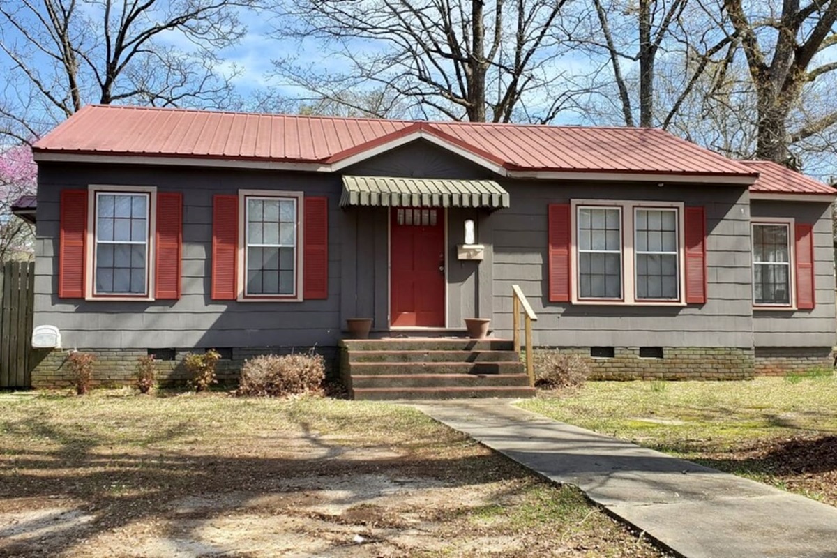 Charming single-story home with a classic red door, cozy front porch, and spacious yard shaded by mature trees.