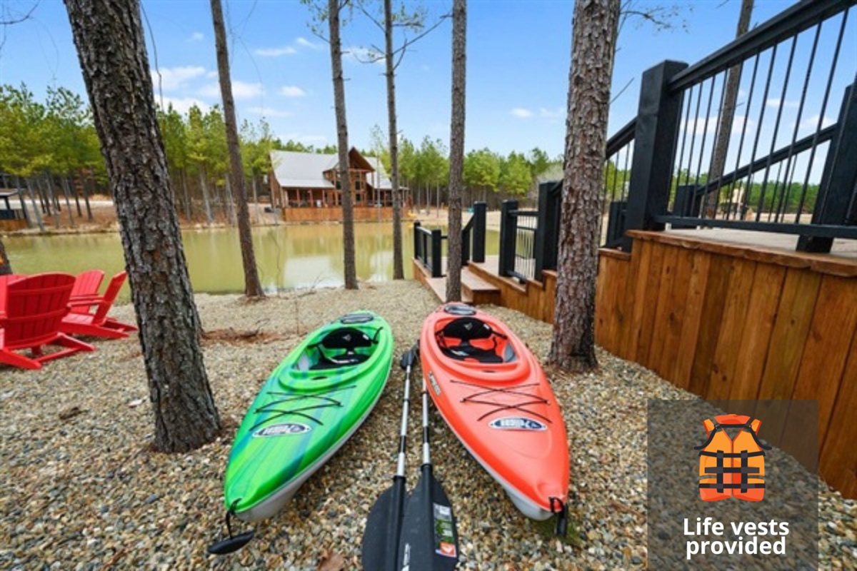 Two colorful kayaks rest on a gravel shore next to a tranquil lake, surrounded by tall pine trees. The setup suggests a peaceful getaway with easy access to water activities.