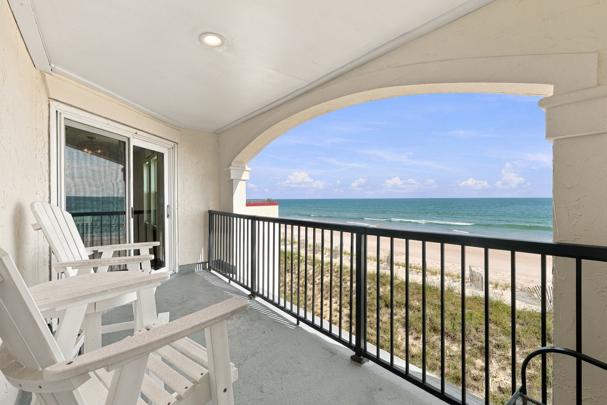Covered oceanfront balcony featuring crisp white Adirondack chairs, an arched overhead architecture detail, sleek black railing, and sweeping views of the sandy beach and turquoise horizon.   