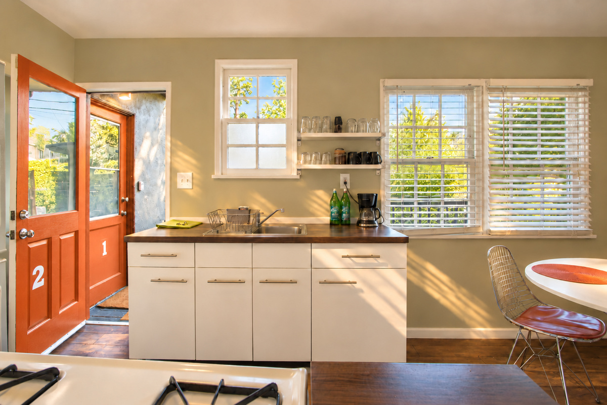 Another kitchen perspective shows white cabinets, the wood countertop and stainless-steel sink beneath the windows. Open shelves keep glassware close at hand, with the round dining table to the right and the orange exterior doors visible to the left.