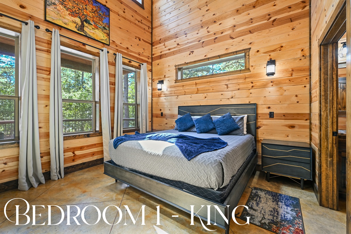 Bedroom 1 features a king bed, high ceilings, and large windows that invite in natural light and peaceful forest views.