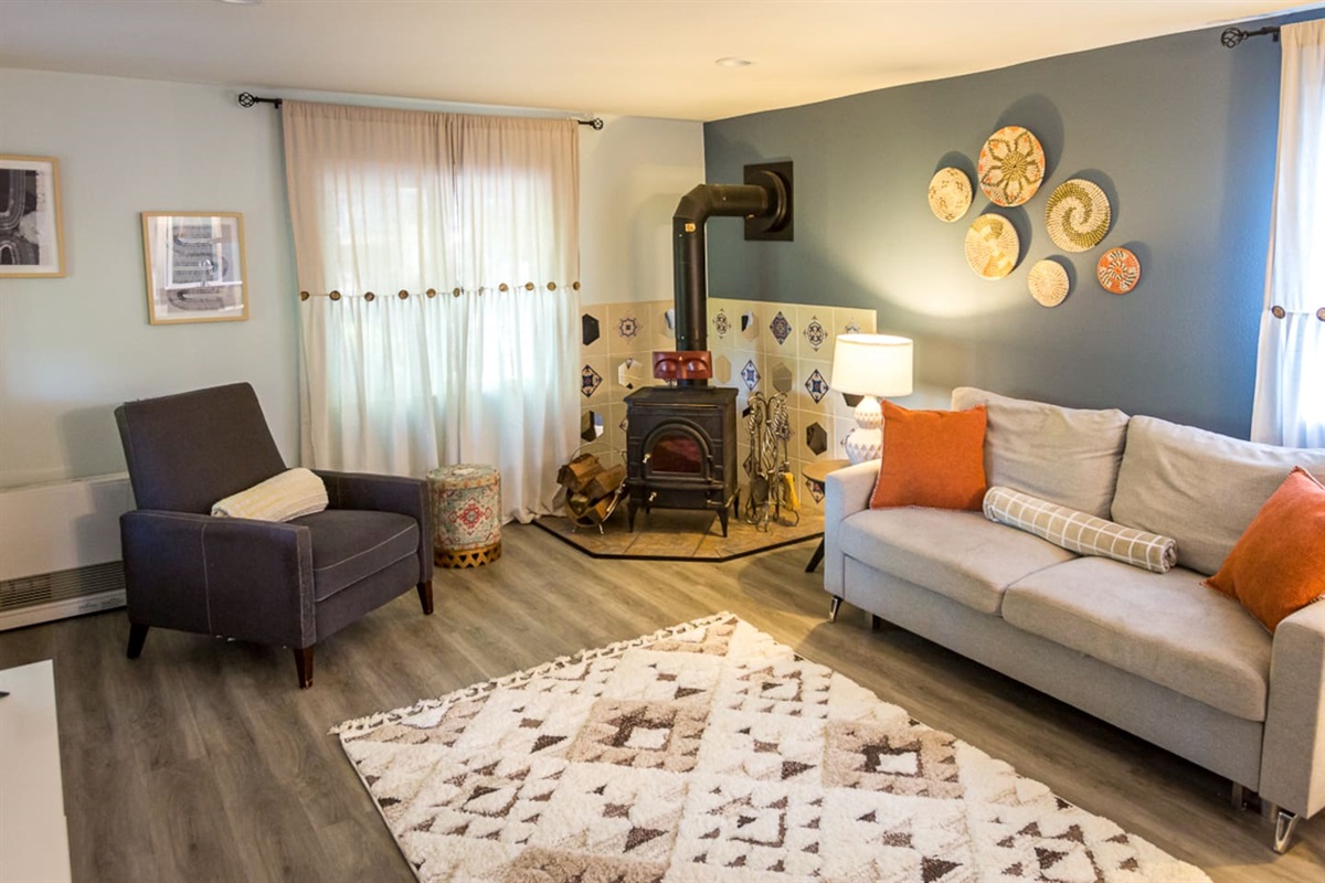 - Welcoming living room with wood burning stove framed by decorative tile surround  
- Plush sofa & accent pillows create a warm & inviting spot to unwind & relax  
- Sunlight filters through soft curtains, adding to the room’s cozy charm & appeal  
