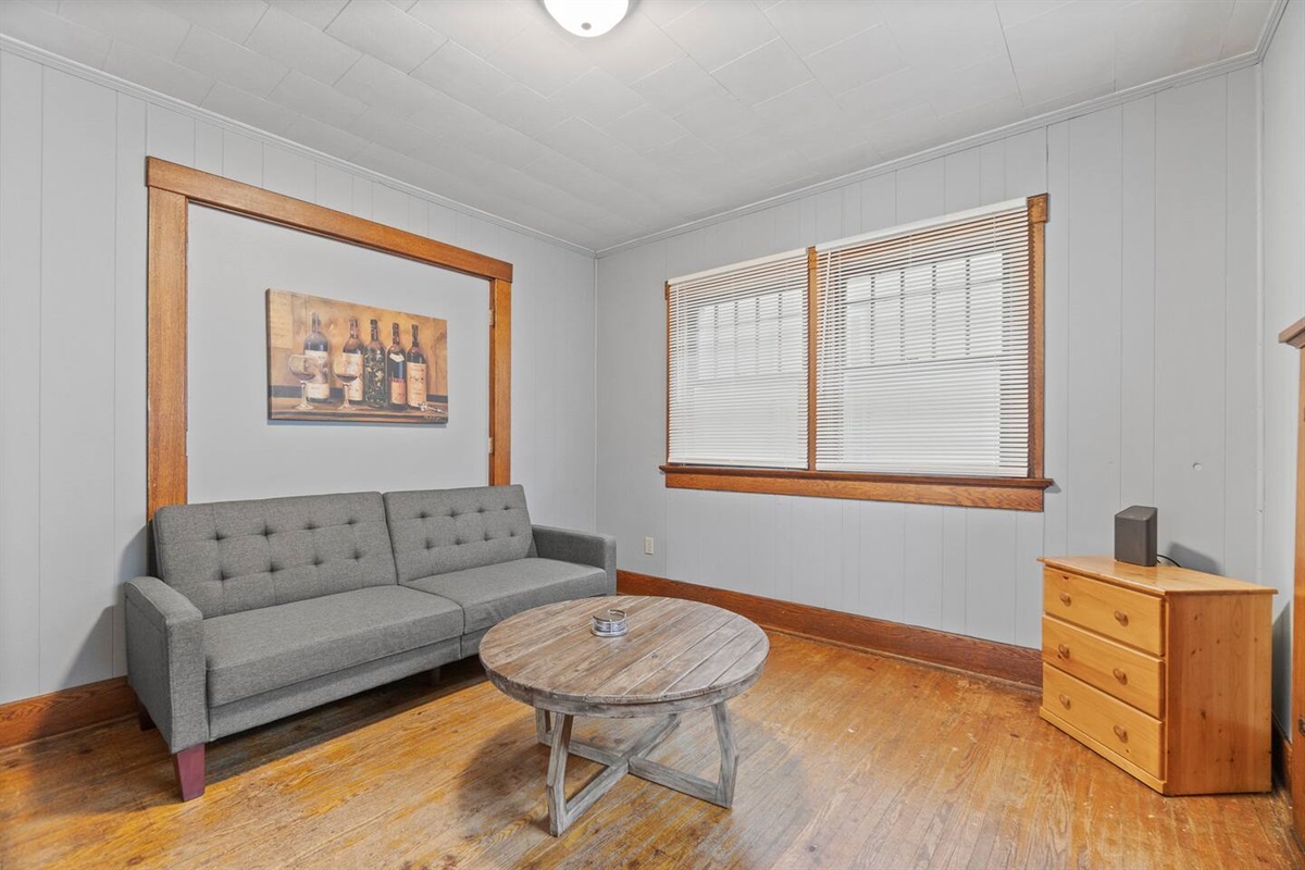 Cozy living room featuring a comfortable bed, charming decorations, large windows for natural light, a tiny table in the middle, a drawer in the corner, and sleek wood flooring—blending comfort and style in a welcoming, relaxed space