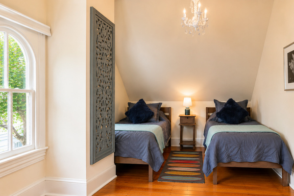 The third floor twin bedroom — tucked under the eaves with a crystal chandelier, original hardwood floors, and arched windows that look out into the treetops.