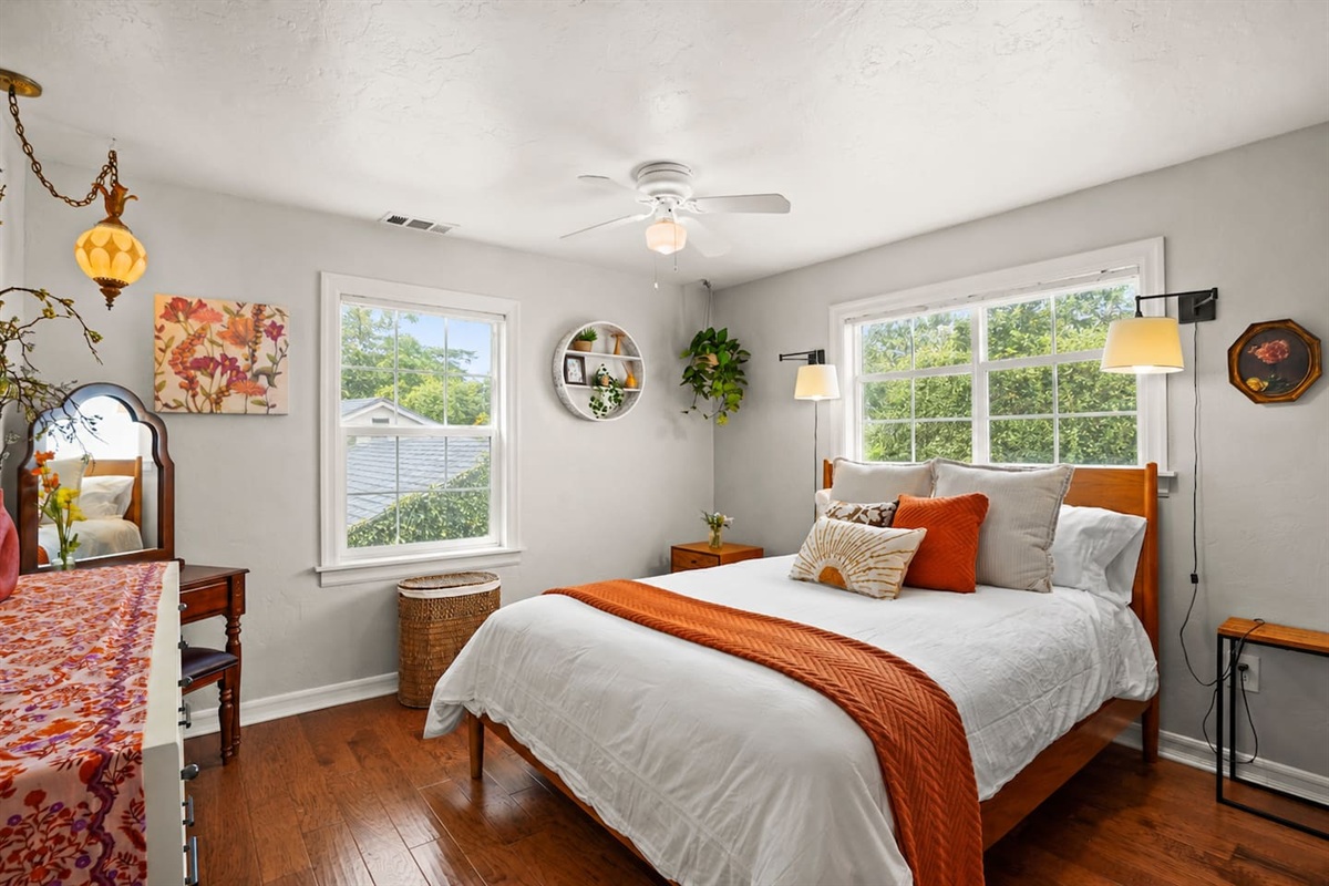 Bright and comfortable main bedroom with a queen bed, dresser, closet, vanity table, and laundry basket.