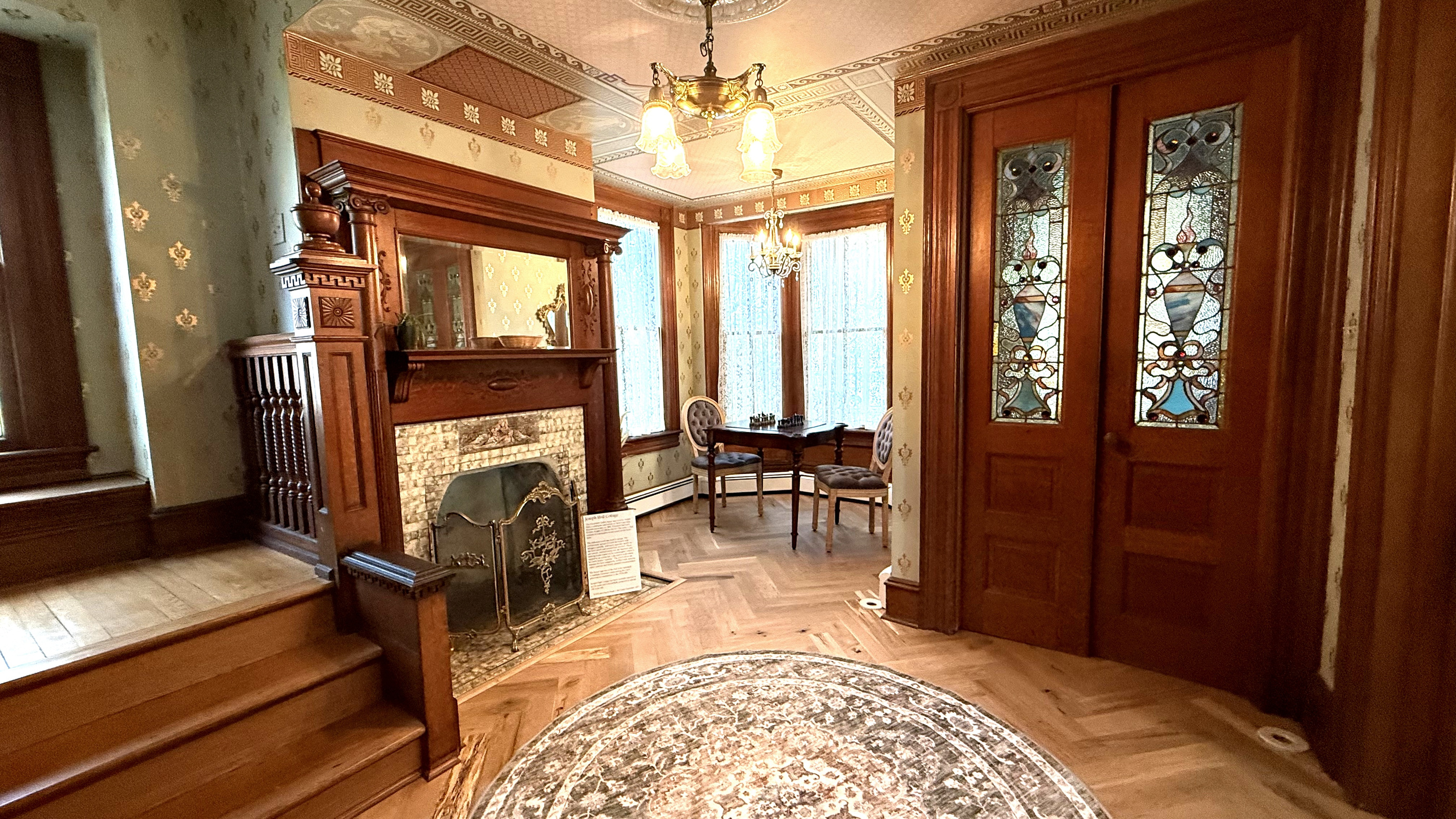 The main foyer — original Victorian wallpaper, a carved walnut fireplace, herringbone hardwood floors, and stained glass entry doors. This is the room that tells you exactly where you are.
