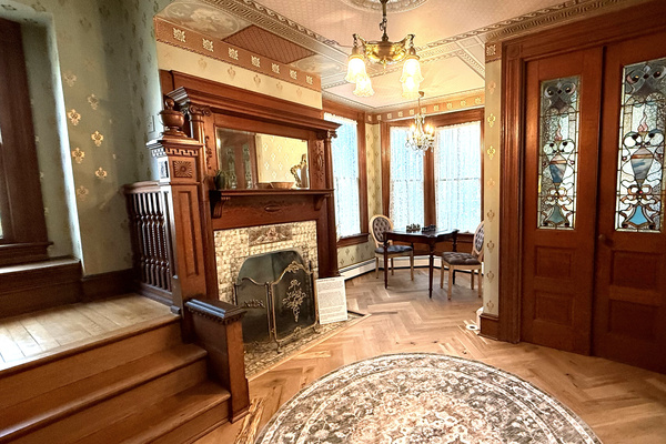 The main foyer — original Victorian wallpaper, a carved walnut fireplace, herringbone hardwood floors, and stained glass entry doors. This is the room that tells you exactly where you are.