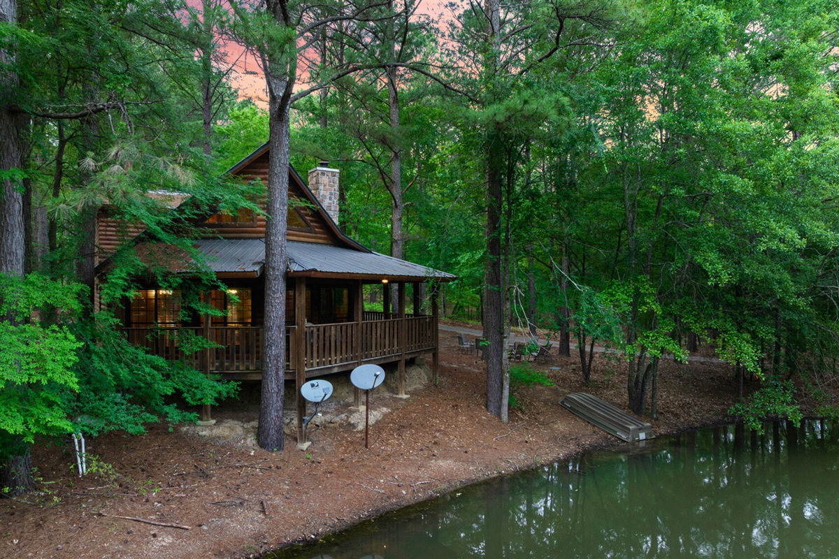 Cabin side view with pond and forest surroundings.
