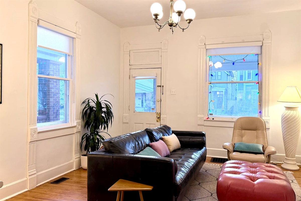A bright, well-scaled living room framed by tall windows and original architectural details, with direct access to the large covered front porch.