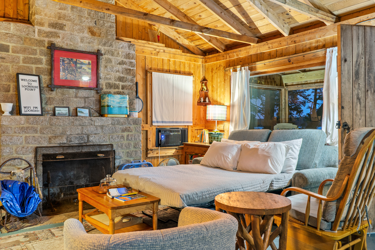 Living room converted into sleeping space at Loonrise Point! Sleeper sofa unfolds into a cozy bed beside the window AC unit and large picture window. Flexible sleeping option inside our authentic, uninsulated seasonal camp in Otis, ME.