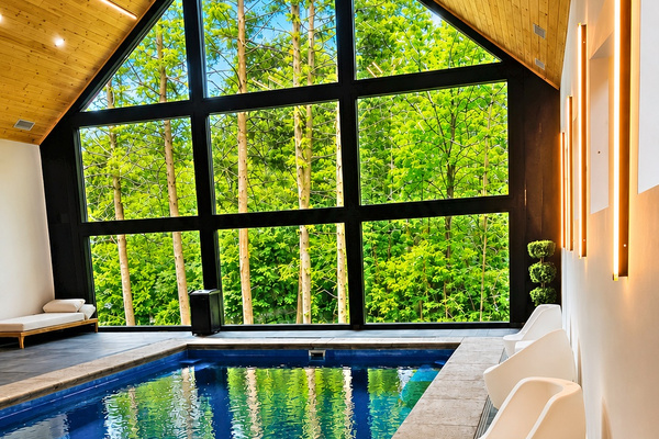 Indoor pool: A-frame window wall onto the forest, white lounge chairs poolside. Swim with a view of the trees.