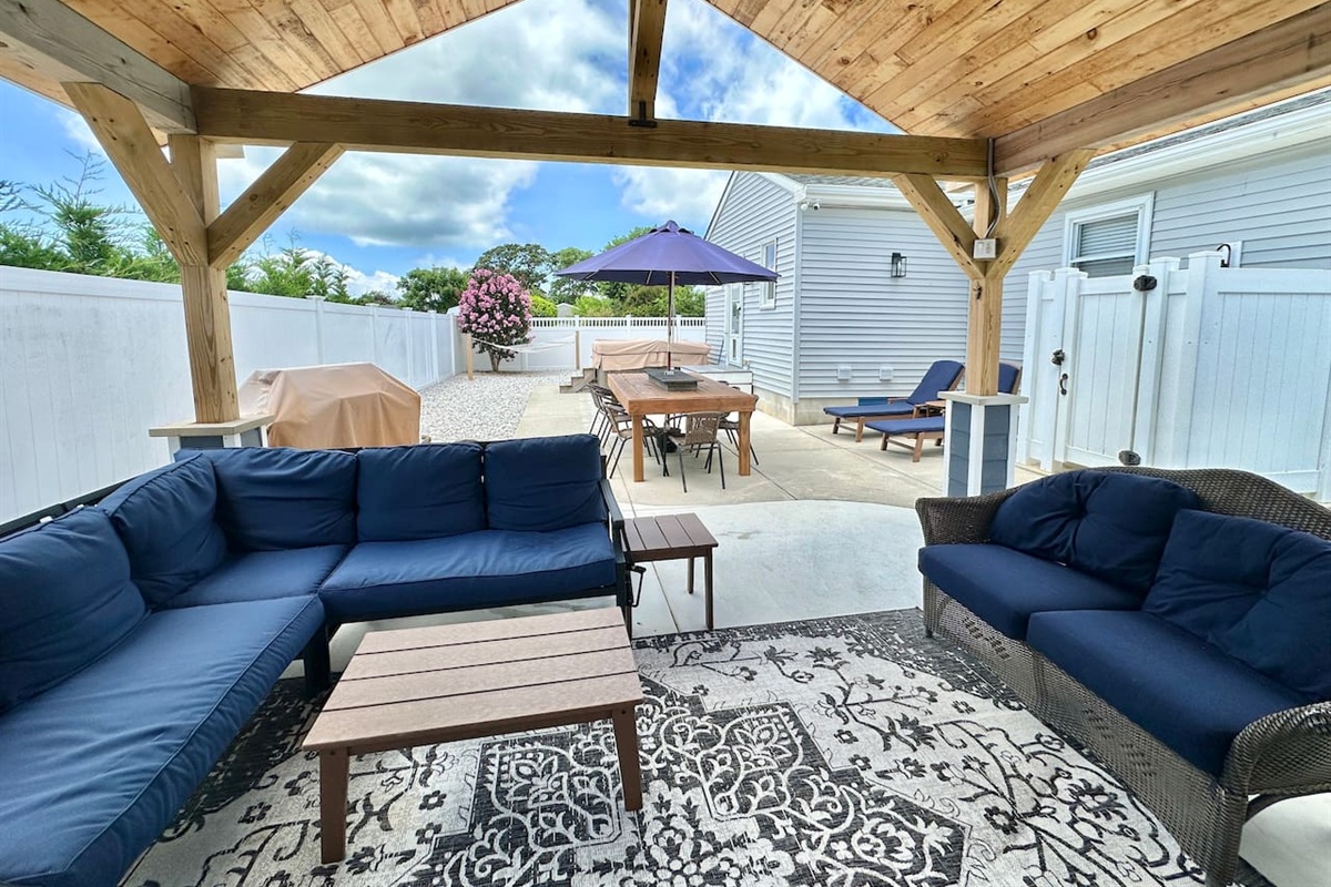 Relax under this wooden pavilion featuring navy sectional seating and a patterned rug. The space offers views of the hot tub, outdoor dining area, and grill, providing a perfect setting for all-day entertainment.