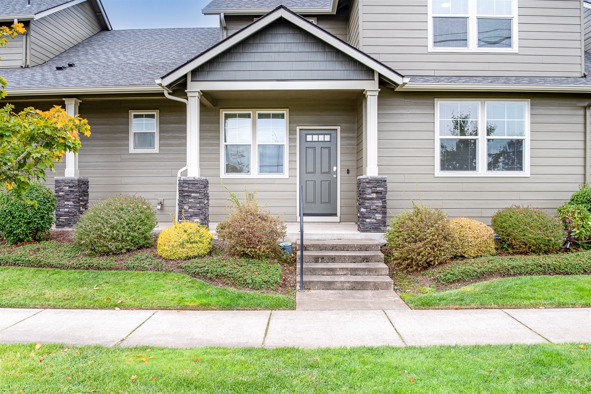 A modern, two-story home featuring a dark gray front door with decorative windows, flanked by stone column accents, and surrounded by neatly landscaped greenery and a concrete pathway leading to the entrance.