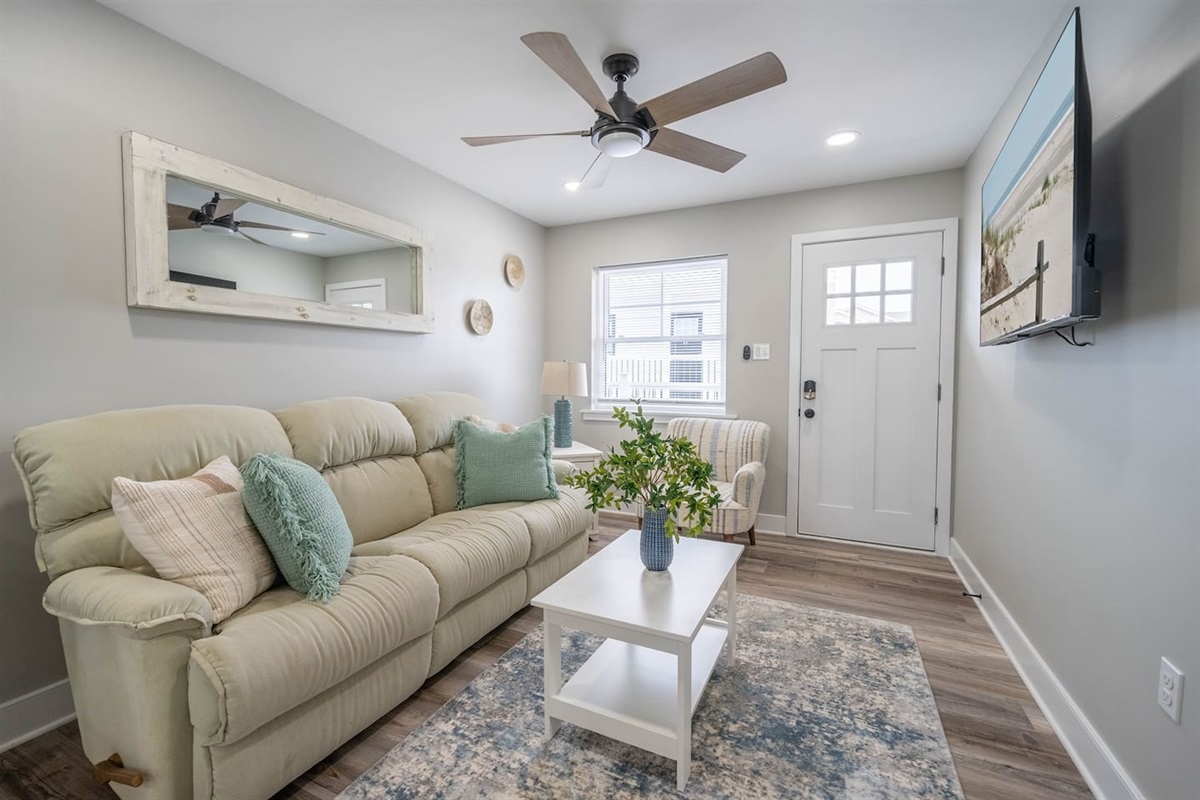 Bright, inviting living area with plush seating, ceiling fan, and wall mounted TV. Natural light and soft coastal accents create a relaxed space to unwind, enjoy a movie, or ease into the day after exploring nearby attractions.