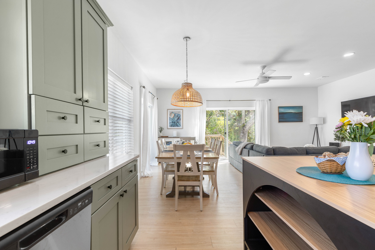 Look down the line from the kitchen: island, table, sofa, then the deck doors and the trees past them. Hand a plate along and it reaches the table without anybody having to take more than a couple of steps.
