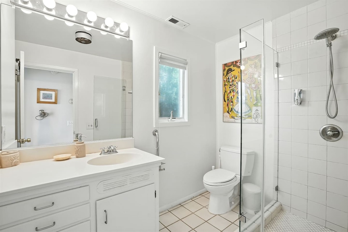 - Modern white bathroom with glass enclosed shower and tile flooring  
- Features vanity with large mirror, bright lighting, and plenty of counter space  
- Window allows soft daylight for a fresh, airy ambiance