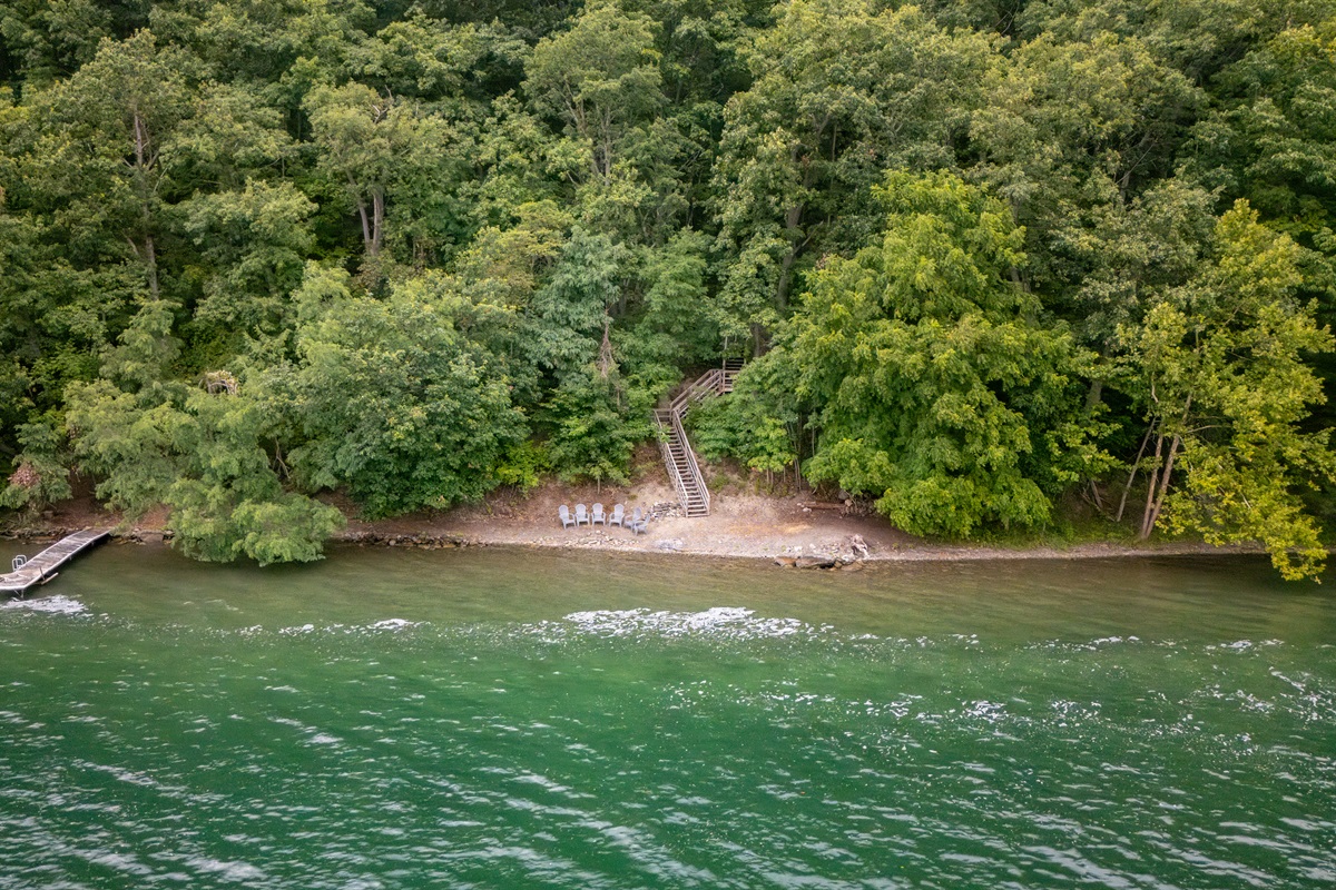 Aerial view of a private, wooded shoreline retreat with direct lake access, dock, and Adirondack chairs for lakeside relaxation.